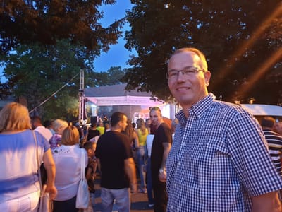 Mann mit Brille bei einem abendlichen Fest unter Bäumen, hinter ihm viele Gäste